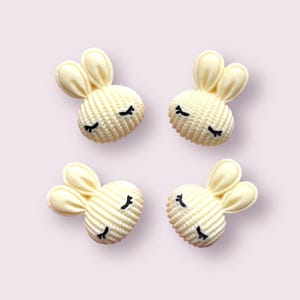 May include: Four cream-coloured bunny head embellishments with black accents. Each bunny has two upright ears and closed eyes. The bunny heads have a textured, woven appearance, and are arranged on a light pink background.