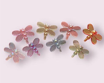 May include: Assortment of eight iridescent dragonfly-shaped embellishments in various colors, including pink, orange, gold, and silver. Each dragonfly has a beaded body and intricate wing details.
