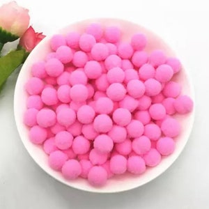 May include: A white bowl filled with numerous small, fluffy, pink pom-poms. The pom-poms are densely packed, creating a textured surface. A few green leaves and a pink flower are visible in the background.