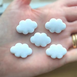 May include: Five small, white cloud-shaped embellishments. Each cloud has a puffy, rounded shape with a slightly textured surface. The clouds are arranged on a light-colored background.