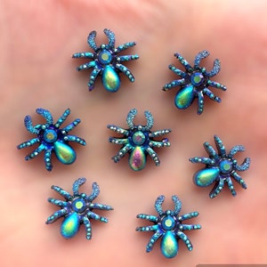 May include: Seven iridescent blue spider-shaped embellishments with crystal accents. Each spider has a rounded body and eight legs, with a large crystal at the centre. The spiders are arranged on a light pink surface.