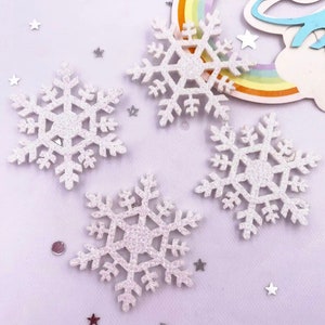 May include: Four white glitter snowflake decorations on a light purple background. The snowflakes are made of foam and have a sparkly finish.