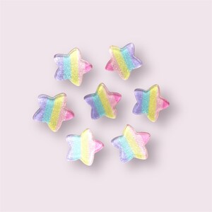 May include: Eight pastel rainbow star-shaped sweets on a light pink background. The sweets are made of a clear, translucent material with a glittery finish.