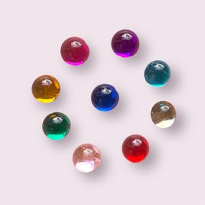May include: Nine colourful glass marbles in various colours including red, pink, blue, green, and gold. The marbles are arranged in a circle on a light purple background.