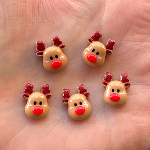 May include: Five small reindeer-shaped ornaments. Each has a tan face, red nose, black eyes, and red antlers. These ornaments are likely for festive decorations or craft projects.