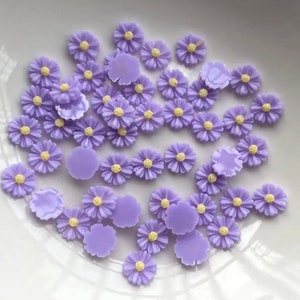 May include: A collection of purple resin daisy flower embellishments. The flowers have a yellow centre and are perfect for adding a touch of whimsy to crafts.