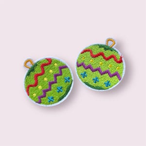 May include: Two embroidered patches in the shape of Christmas ornaments. The ornaments are green with red, purple, and blue decorative stitching. Each ornament has a gold loop at the top.