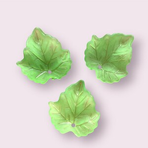 May include: Three light green leaf-shaped decorative items with a hole in each. The leaves have detailed veining and are arranged on a light pink background. The leaves appear to be made of glass or a similar translucent material.