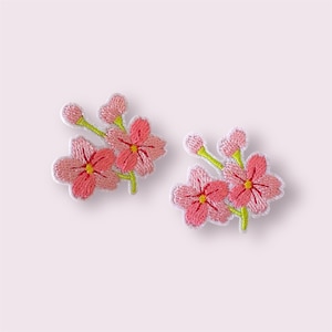 May include: Two embroidered floral patches featuring pink cherry blossoms with yellow centers and green stems. The flowers have a white outline. These patches are ideal for adding a decorative touch to clothing or accessories.