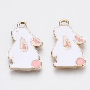 May include: Two gold-coloured metal charms shaped like white rabbits with pink ears and feet. The charms have a loop at the top for attaching to jewellery or other crafts.