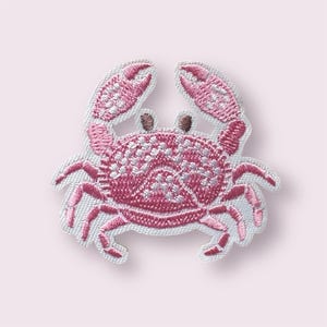 May include: Embroidered patch of a pink crab with white detailing. The crab's claws, legs, and body are intricately stitched. The patch is set against a light pink backdrop, creating a visually appealing design. The patch is approximately 7 cm wide.