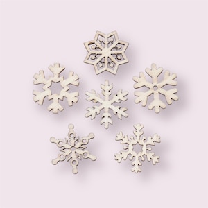 May include: Five wooden snowflakes in various designs are arranged on a light pink background. The snowflakes are cut from light-colored wood and feature intricate patterns. These decorative pieces are ideal for winter-themed crafts and holiday decorations.