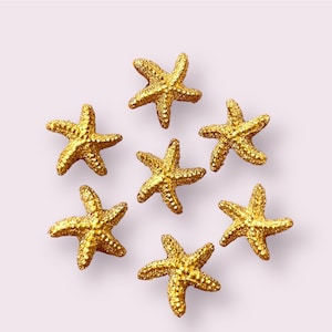May include: Seven gold starfish arranged in a circle on a light pink background.