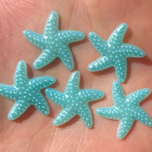 May include: Five light blue starfish with a pearlescent finish. The starfish are arranged in a cluster on a light pink surface.