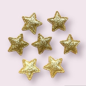 May include: Seven gold glitter star-shaped embellishments on a light pink background.