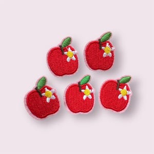 May include: Five embroidered apple patches. Each patch features a red apple with a green leaf and a small white flower with a yellow centre. The edges of the apples are outlined in a light pink colour.