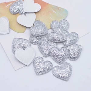 May include: A pile of silver glitter heart-shaped embellishments. The hearts are puffy and have a raised edge.