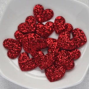 May include: A collection of small, red glitter heart-shaped embellishments. The hearts are scattered in a white, shallow dish. The glitter gives them a sparkling appearance, and they vary slightly in size and shape.