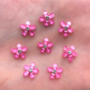 May include: Eight pink flower-shaped embellishments with a clear rhinestone center. Each flower has five petals and a white outline.