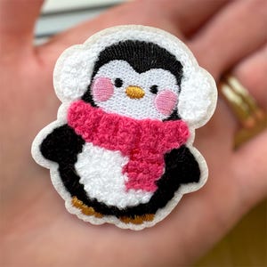 May include: Embroidered penguin patch. The penguin has a black and white body, a pink scarf, and white earmuffs. It features a yellow beak and pink cheeks, outlined in white.