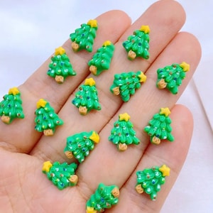 May include: A collection of 15 green Christmas tree shaped resin embellishments with gold star toppers. The embellishments are scattered on a hand.