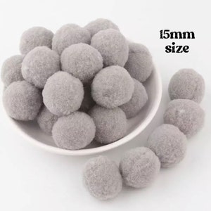 May include: A collection of grey pom-poms, each approximately 1.5cm in size, are displayed in a white bowl and scattered around it. The pom-poms have a soft, fuzzy texture. The text "15mm size" is visible in the image.