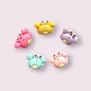 May include: Five colourful plastic crab figurines with smiling faces. The crabs are in various colours, including yellow, pink, purple, light blue and peach.