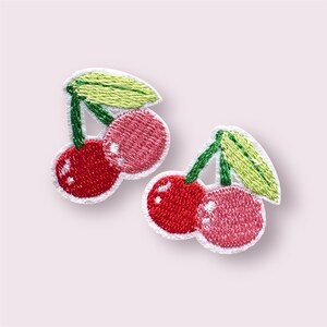 May include: Two embroidered cherry patches. Each patch features two cherries in shades of red and pink, with green stems and leaves. The patches have a white border and are set against a soft pink background.