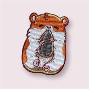 May include: Embroidered patch featuring a cartoon hamster in shades of orange, white, and brown. The hamster is holding a large sunflower seed. The patch has a detailed, stitched design with a light pink background.
