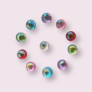 May include: A circular arrangement of iridescent glass beads in shades of blue, green, red, and purple. The spherical beads have a textured surface and are set against a pale pink backdrop. Each bead is approximately 1.5 cm in diameter.
