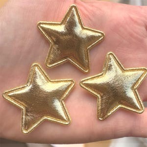 May include: Three gold star-shaped patches with a metallic sheen. Each star has a stitched border, and they are arranged in a close-up shot. These decorative patches are suitable for various craft projects.