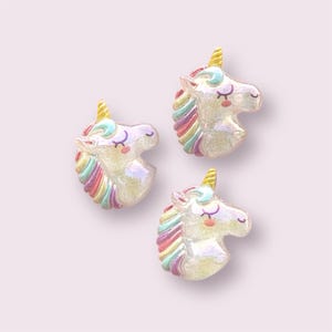 May include: Three iridescent white unicorn head charms with rainbow manes and gold horns. The unicorn heads are facing right and have closed eyes and pink cheeks.