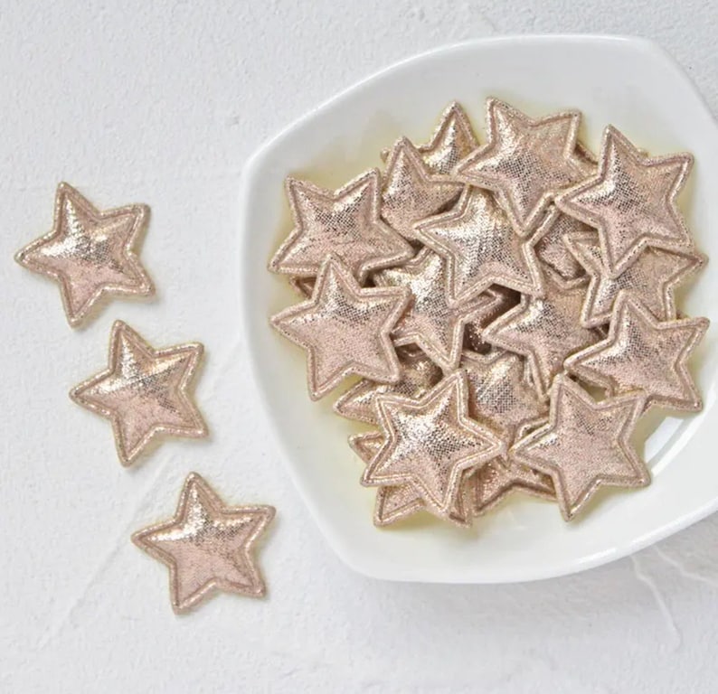 Pode incluir: Uma tigela branca cheia de enfeites em forma de estrela dourados com glitter. As estrelas est&atilde;o espalhadas pela tigela e algumas se sobrep&otilde;em.