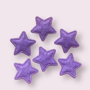 May include: Six purple felt stars with a glittery finish. The stars are arranged in a semi-circle on a white background.