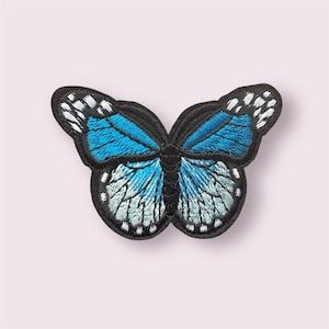 May include: Embroidered butterfly patch featuring a detailed design with shades of blue and white on a black background. The butterfly has a symmetrical wing pattern and is suitable for clothing or accessories.