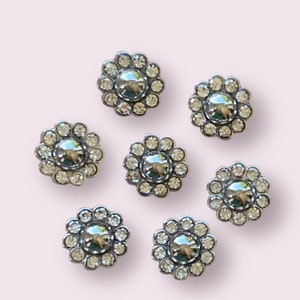 May include: Seven silver-toned flower-shaped buttons with clear rhinestones. Each button has a round, silver-toned center.
