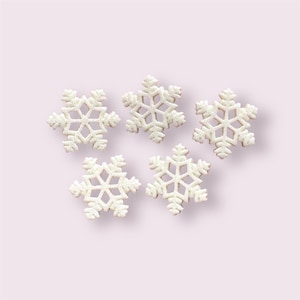 Peut inclure: Cinq décorations blanches en forme de flocons de neige avec des paillettes sont disposées sur un fond rose clair. Les flocons de neige présentent un design ajouré et délicat, idéal pour les décorations de vacances d'hiver.