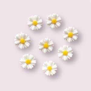 May include: Eight white daisy flower embellishments with yellow centres. The daisies are arranged in a circle on a light pink background.