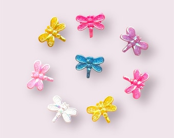 May include: A collection of eight colorful dragonfly-shaped embellishments. The dragonflies are in various shades, including yellow, pink, blue, and iridescent white. Each dragonfly has a glittery finish, adding a touch of sparkle. They are arranged in a circular pattern against a soft pink background.