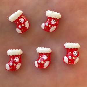 May include: Five miniature Christmas stockings in red with white trim and snowflake designs. The stockings have white cuffs and heels, and small yellow dots are scattered across the red surface. The background is a solid, muted pink.