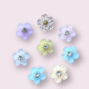 May include: Assortment of eight flower-shaped embellishments with rhinestones in the center. The flowers are in shades of light blue, lavender, yellow, and white.