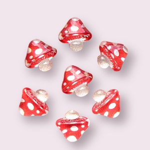 May include: Seven red and white mushroom shaped glass beads with white dots. The beads are arranged in a circle on a white background.