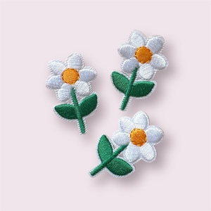 May include: Three embroidered daisy patches. Each patch features white petals, a yellow centre, and green leaves and stem. The patches are arranged on a light pink background.