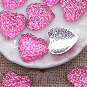 May include: Close-up of several pink heart-shaped embellishments with a glittery texture. One heart has a silver metallic finish. The hearts are arranged on a white, scalloped-edge plate, suggesting a craft or decorative theme.