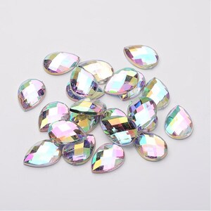 May include: A collection of iridescent, teardrop-shaped rhinestones. Each gem has a faceted surface that reflects a rainbow of colors. The rhinestones are scattered on a white surface, creating a sparkling effect. Ideal for crafting and embellishments.