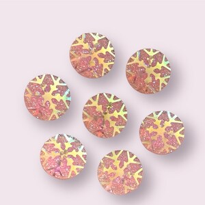 May include: Seven iridescent pink and gold snowflake-shaped embellishments. Each embellishment is a round circle with a snowflake design.