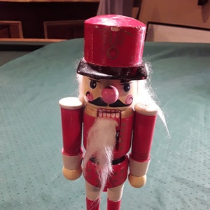 Pode incluir: Uma figura de quebra-nozes de madeira em um design clássico. A figura é principalmente vermelha com um chapéu preto, bigode e botas. Possui barba branca e nariz rosa. O quebra-nozes está em uma base redonda.
