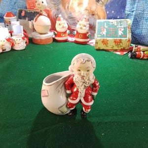 May include: A ceramic Santa Claus figurine with a white beard and red suit, holding a white sack. Other Christmas decorations, including Santa figurines and a gift box, are arranged on a green surface.