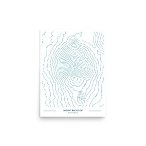 May include: A white print featuring a teal topographic map of Mount Bachelor, with contour lines. The text "MOUNT BACHELOR" is centered at the bottom, with the elevation in feet below.
