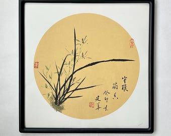 May include: A framed print of a traditional Chinese ink painting depicting a stylised orchid plant with delicate flowers and leaves. The painting is set against a circular background of pale yellow and features a calligraphic inscription in black ink.